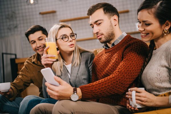 cheerful group of friends looking at smartphone while holding drinks