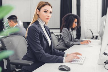 Masaüstü bilgisayar kullanarak ve kamera Office bakarak güzel genç iş kadını 