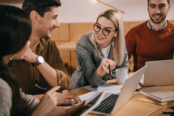 улыбающаяся женщина в очках, улыбающаяся возле ноутбука, разговаривая с коллегами
