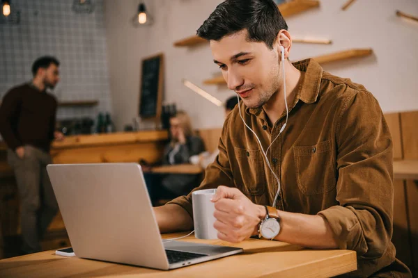 handsome man in earphones holding cup of coffee and looking at laptop in cafe