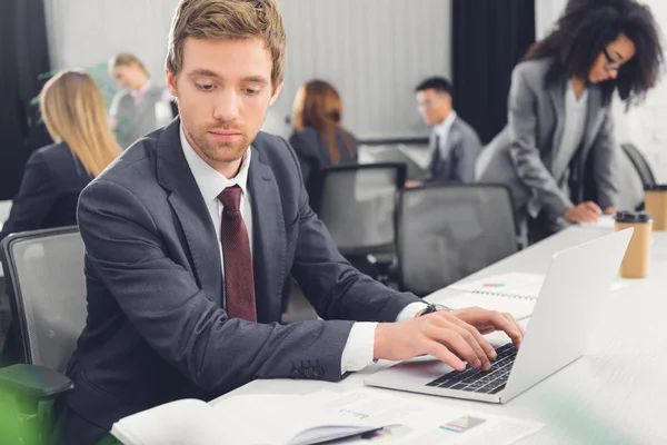 Seriöser Junger Geschäftsmann Schaut Auf Notizbuch Und Benutzt Laptop Büro — Stockfoto