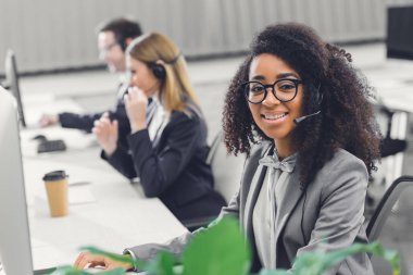 çekici genç Afro-Amerikan kadın ofiste meslektaşları ile çalışırken kameraya gülümseyen kulaklık 