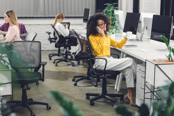 非洲裔美国女商人拿着杯子 用智能手机说话 同时与同事在办公室工作 — 图库照片