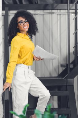 kağıtları tutarak ve kamera Office gülümseyen çekici genç Afro-Amerikan işkadını 