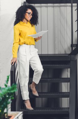kağıtları tutarak ve Office merdivenlerde durup güzel gülümseyen genç Afro-Amerikan işkadını 