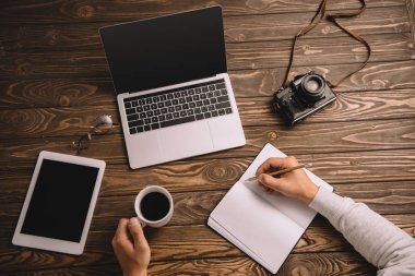 Erkek freelancer defter masada fincan kahve, dizüstü bilgisayar, tablet ve retro fotoğraf makinesi ile yazma görünümünü kırpılmış