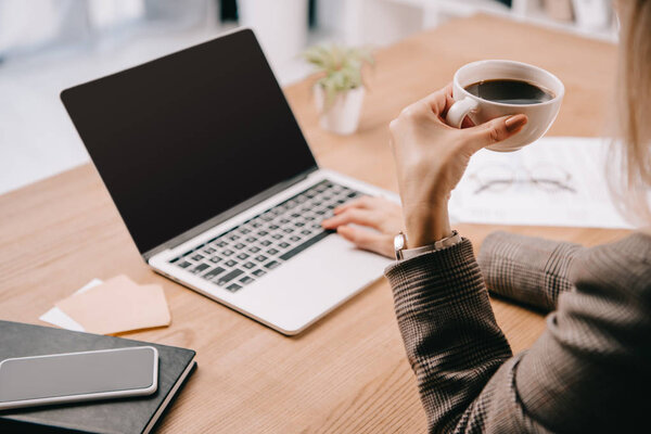 cropped view of businesswoman holding cup of coffee while working with laptop in office