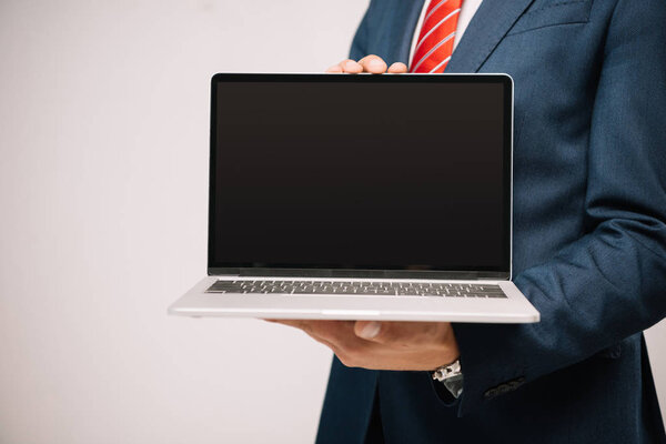 cropped view of businessman in suit presenting laptop with blank screen isolated on grey