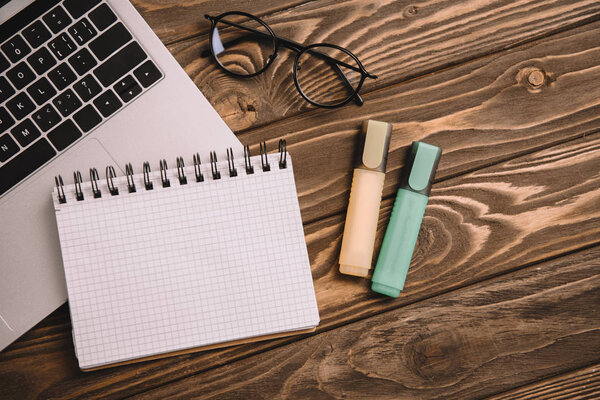 top view of laptop, empty notebook, markers and eyeglasses on wooden table