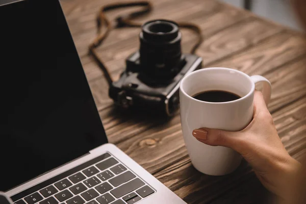 裁剪视图的自由职业者举行一杯咖啡在工作场所与老式的照片摄像头和笔记本电脑 — 图库照片