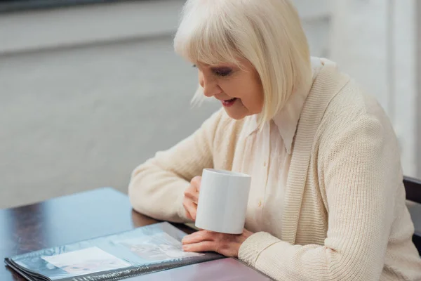 Mujer Mayor Sentada Mesa Mirando Álbum Fotos Mientras Bebe Café — Foto de Stock