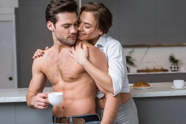 Mulher Atraente Abraçando Homem Sem Camisa Com Xícara Café Cozinha — Fotografia de Stock