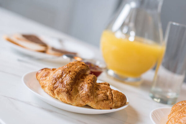 selective focus of delicious croissant on plate with orange juice in jug on background