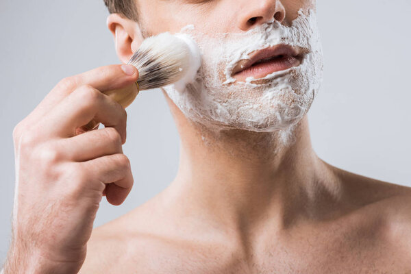 cropped view of young man applying shaving foam with brush, isolated on grey