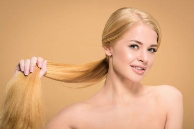 beautiful naked blonde woman holding long hair and smiling at camera isolated on beige 
