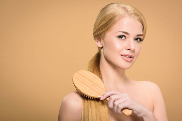 attractive blonde woman brushing smooth hair with wooden hairbrush and smiling at camera isolated on beige 