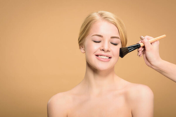 cropped shot of stylist applying makeup with cosmetic brush to happy naked girl isolated on beige  