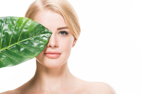attractive naked girl with green tropical leaf near eye looking at camera isolated on white