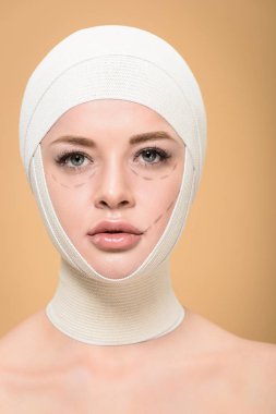 young woman with bandages over head and lines on face looking at camera isolated on beige