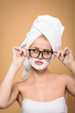 beautiful girl with towel on head and shaving cream on face adjusting eyeglasses and looking away isolated on beige