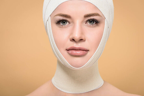 young woman with bandages over head looking at camera isolated on beige, plastic surgery concept