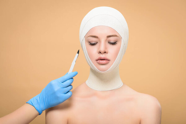 cropped shot of hand in latex glove holding scalpel near woman with bandages over head looking down isolated on beige  