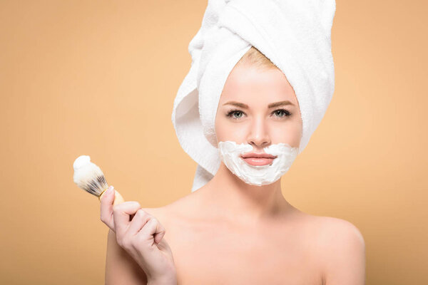 naked woman with towel on head and shaving cream on face holding shaving brush and looking at camera isolated on beige