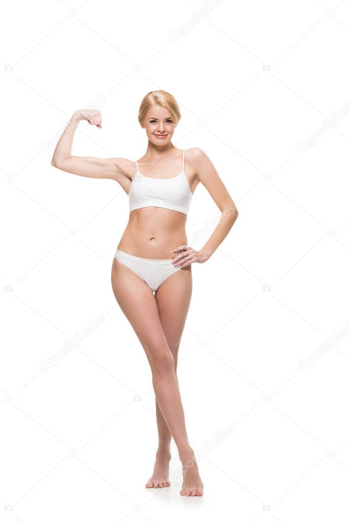 smiling slim girl in underwear standing with hand on waist and showing biceps isolated on white