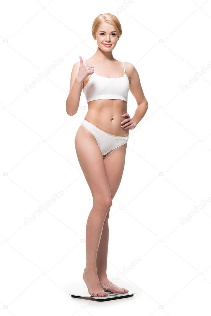 attractive happy slim girl in underwear standing on scales and showing thumb up isolated on white