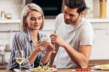 yakışıklı erkek arkadaşı ve çekici kız arkadaşı akıllı telefon kullanarak ve şarap cam tutarak seçici odak