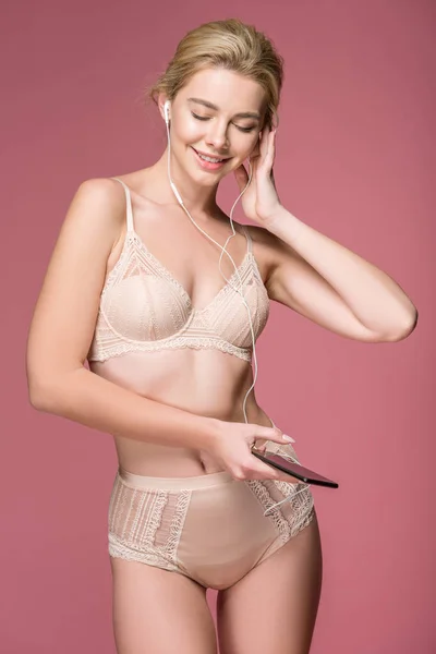 快乐的女人在内衣听音乐与智能手机和耳机 隔离在粉红色 — 图库照片