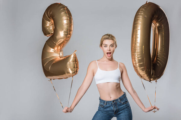 surprised beautiful girl in jeans and white bra posing with 20 golden balloons, isolated on grey
