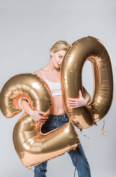 beautiful happy girl in jeans and white bra posing with 20 golden balloons, isolated on grey