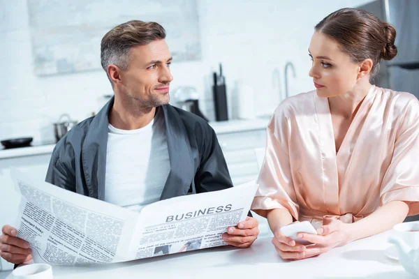 男子在长袍阅读商业报纸 而妇女使用智能手机在早餐在厨房 — 图库照片