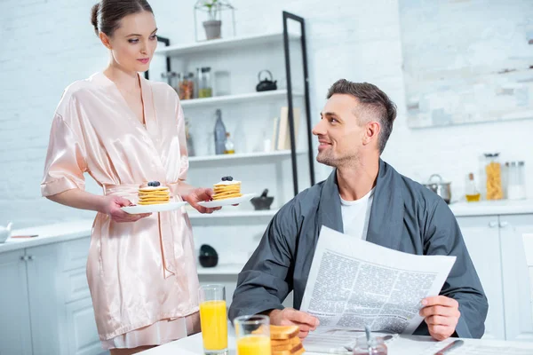 woman in robe holding plates with pancakes while man reading newspaper during breakfast in kitchen