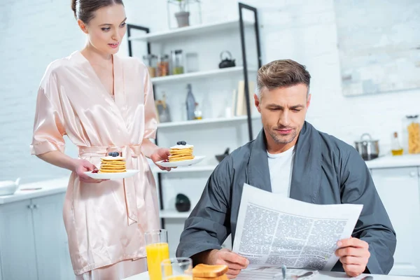 Woman Robe Holding Plates Pancakes While Man Reading Newspaper Breakfast — Stock Photo, Image