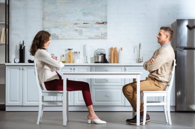 dissatisfied couple looking at each other while sitting with arms crossed at table in kitchen