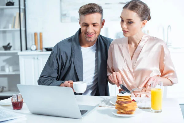 Mutfakta Kahvaltı Yaparken Laptop Ile Masada Oturan Cüppeli Yetişkin Çift — Stok fotoğraf