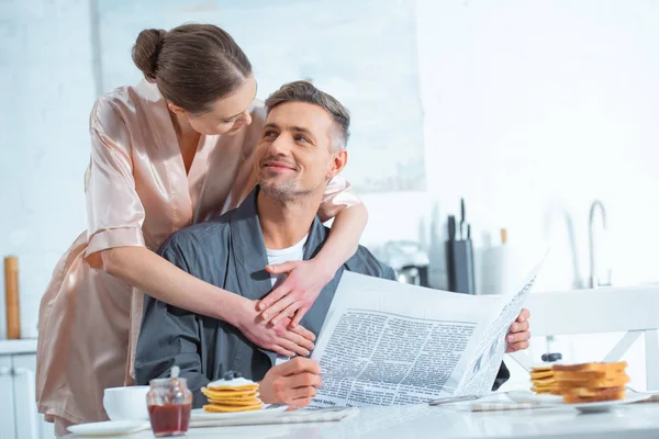 woman in robe embracing handsome man with newspaper during breakfast in kitchen