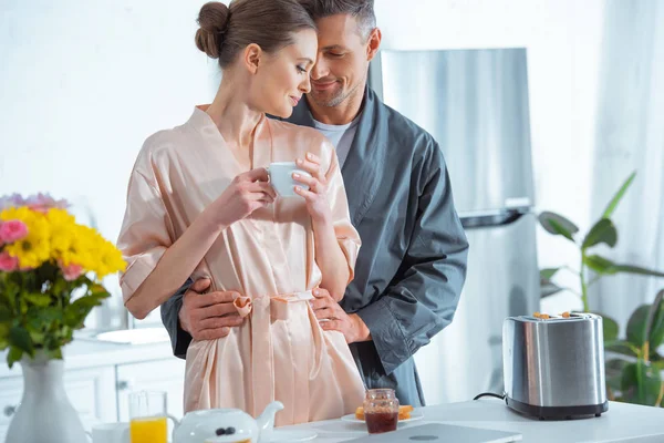 handsome man in robe embracing beautiful woman with cup of tea in kitchen