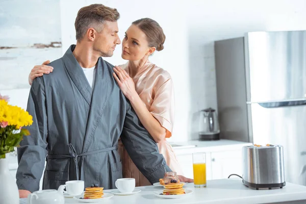 beautiful woman in robe embracing handsome man during breakfast in kitchen