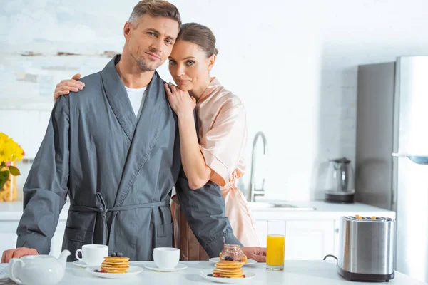 selective focus of beautiful woman in robe embracing man during breakfast in kitchen