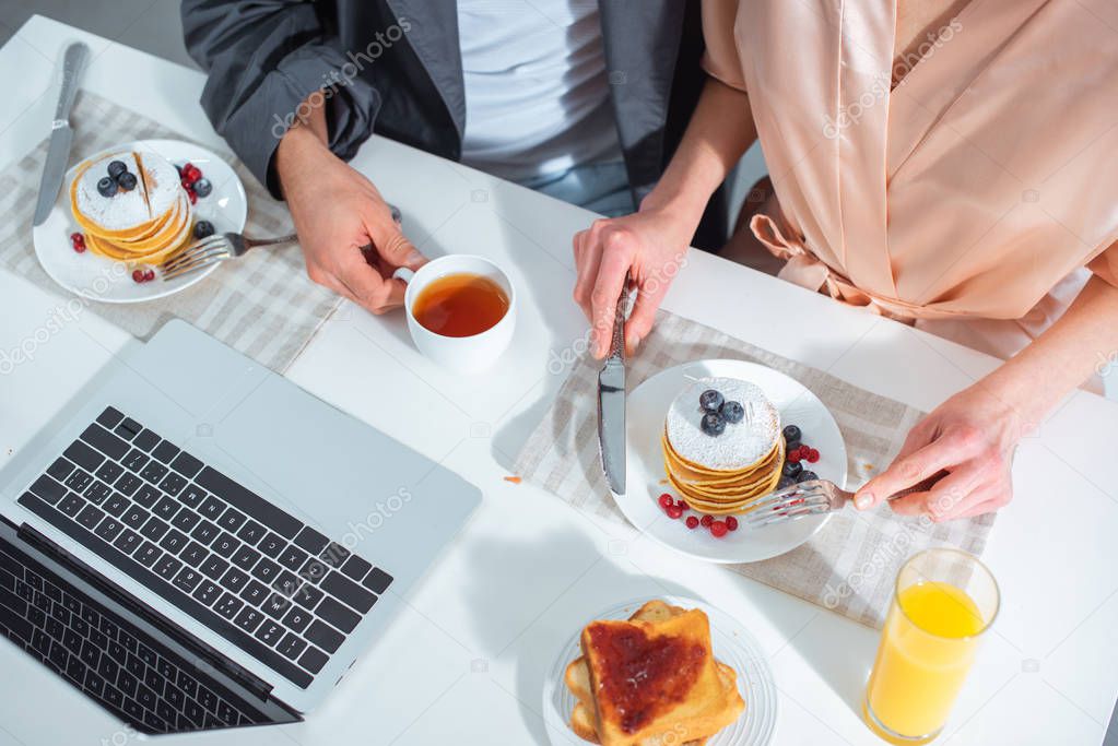 cropped view of adult couple sitting at table with laptop while having breakfast in morning