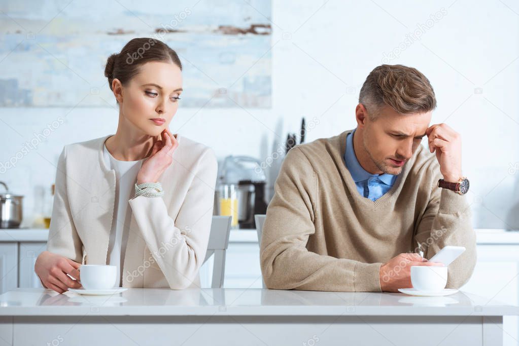 dissatisfied man using smartphone and ignoring woman during breakfast in kitchen