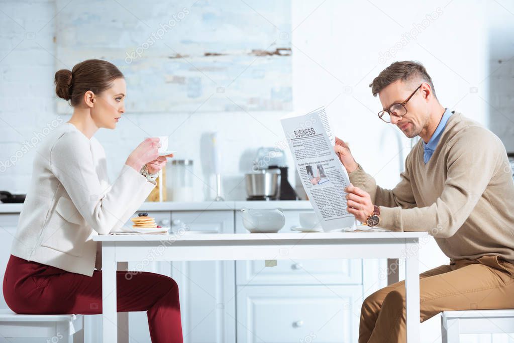 dissatisfied woman drinking tea while man reading business newspaper during breakfast in kitchen