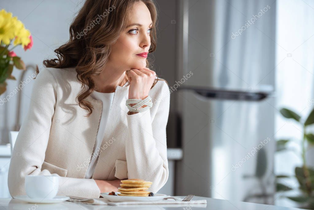 beautiful pensive woman sitting at table with pancakes and coffee during breakfast in kitchen