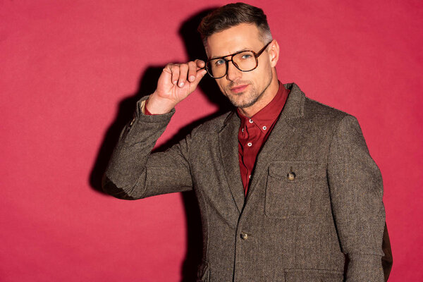 stylish man in formal wear looking at camera and adjusting glasses on red background