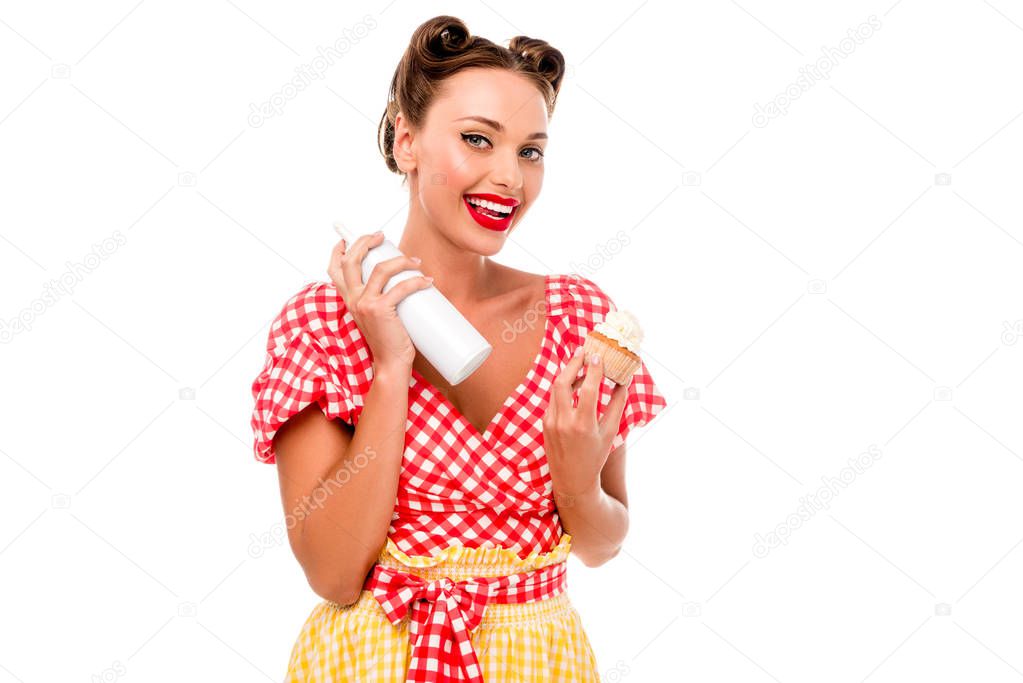 Smiling pin up girl holding cupcake and bottle with whipped cream isolated on white