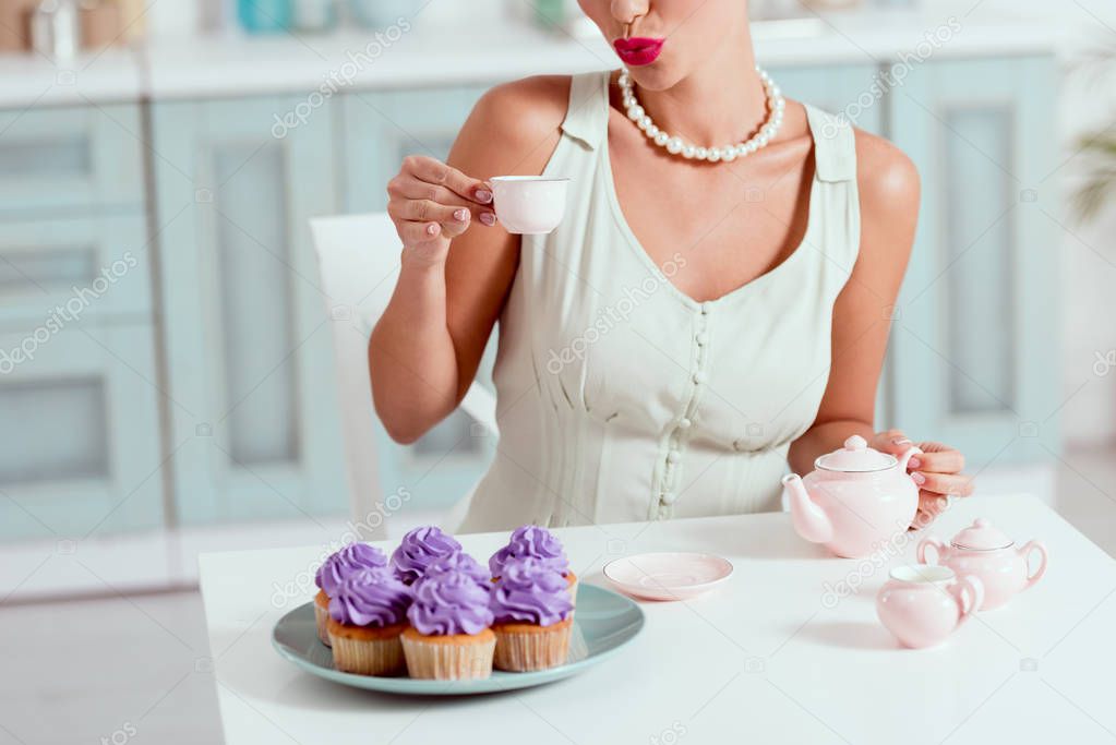 Cropped view of pin up girl holding cup of coffee while sitting at table with plate of cupcakes