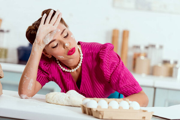 exhausted pin up girl with flour traces on hands and face leaning on kitchen table